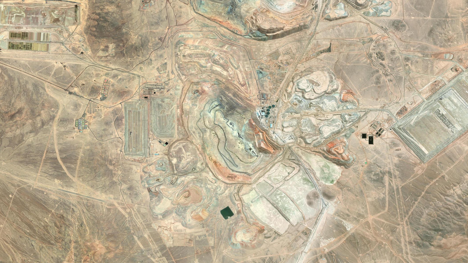 Satellite view of the Escondida copper mine in Chile's Atacama Desert, showing large open pits, concentrator plants, leach pads and tailings deposits.